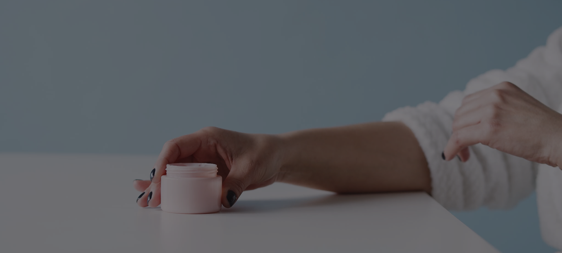 A hand reaching for an open jar of face cream on a pale surface