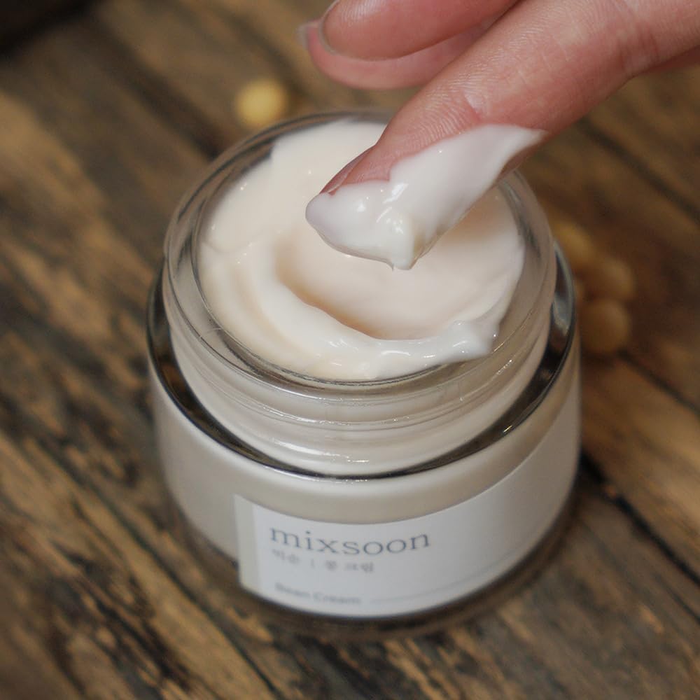 A fingertip scooping thick white cream from the open Bean Cream jar on a wooden surface